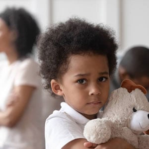 A young boy holds a teddy bear, standing before a group of people.